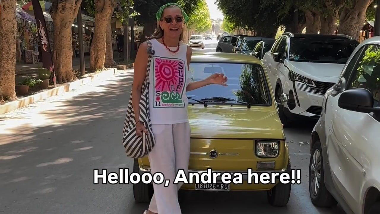 Andrea on a shady market street beside a little yellow Fiat, saying hello